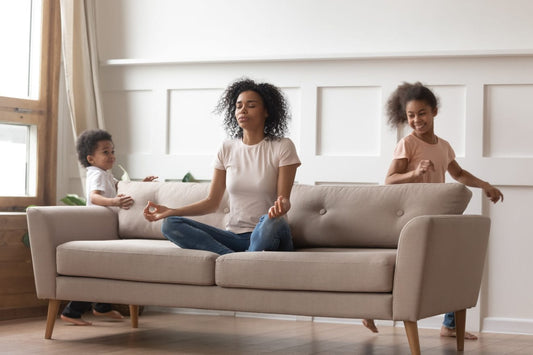 Forælder sidder roligt på sofa og mediterer, mens børn leger omkring, symbol på balance og håndtering af hverdagens stress med små børn